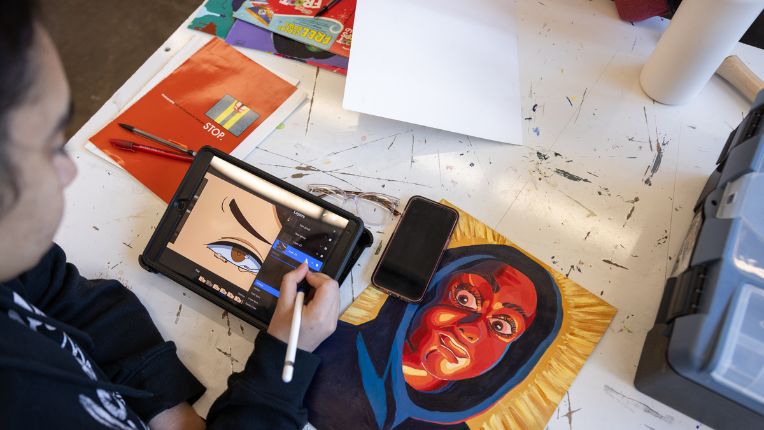 A student is making digital art on an ipad screen while referencing a printed piece of art of a person wearing a hooded sweatshirt.