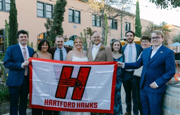 Madeline and Geoff pose with fellow UHart Alumni at their wedding holding a Hartford Hawks flag.