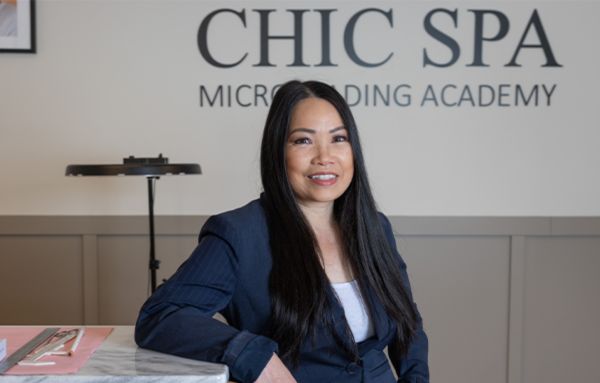 Devanny Do, class of 1998 posing for her headshot in front of her business sign that reads Chic Spa Microblading Academy.